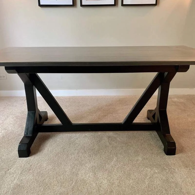 Rectangular wooden table with a light top and dark X-shaped base on carpet, against a wall with three framed pictures.