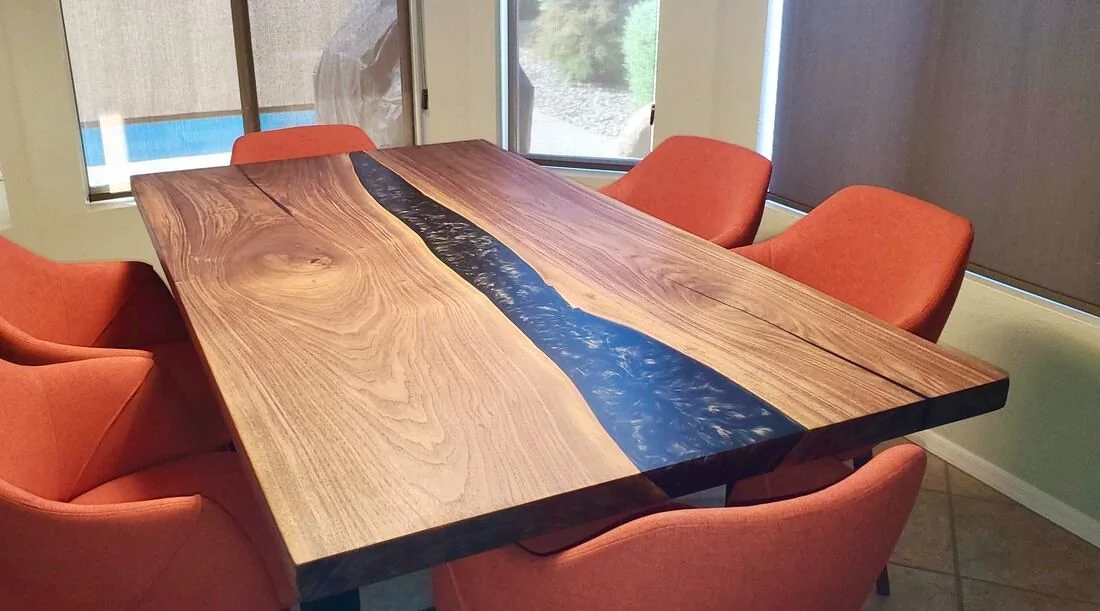 A wooden dining table with a dark blue resin river inlay, surrounded by six orange upholstered chairs in a bright dining area with large windows and blinds.