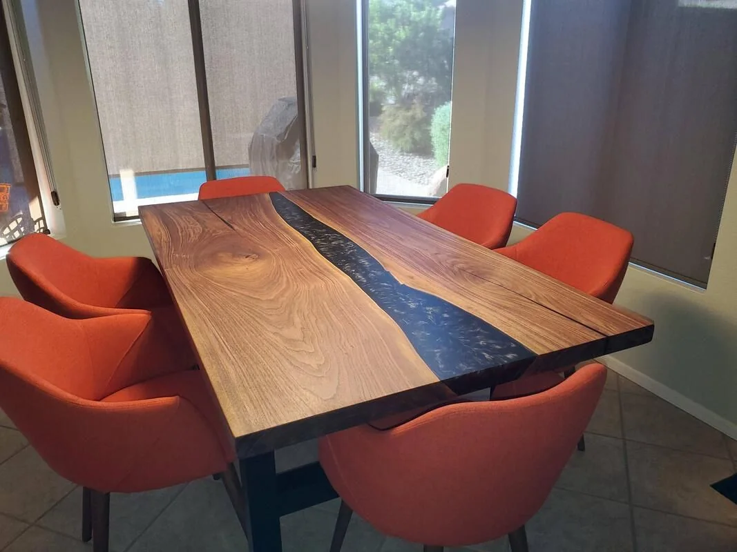 A modern dining area with a rectangular wooden table featuring a central epoxy resin river, surrounded by six orange upholstered chairs, illuminated by natural light from large windows with blinds.
