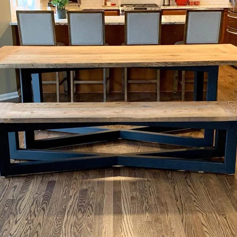 Light wood dining table with matching bench, both featuring dark geometric metal bases, photographed against a dark studio wall.