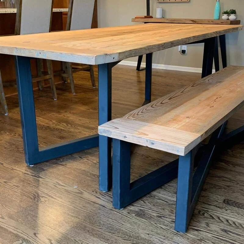 Light wood dining table with matching bench, both featuring dark blue metal frame legs. The set is placed on hardwood flooring in an open-plan kitchen and dining area, with high-backed chairs at a kitchen counter in the background.