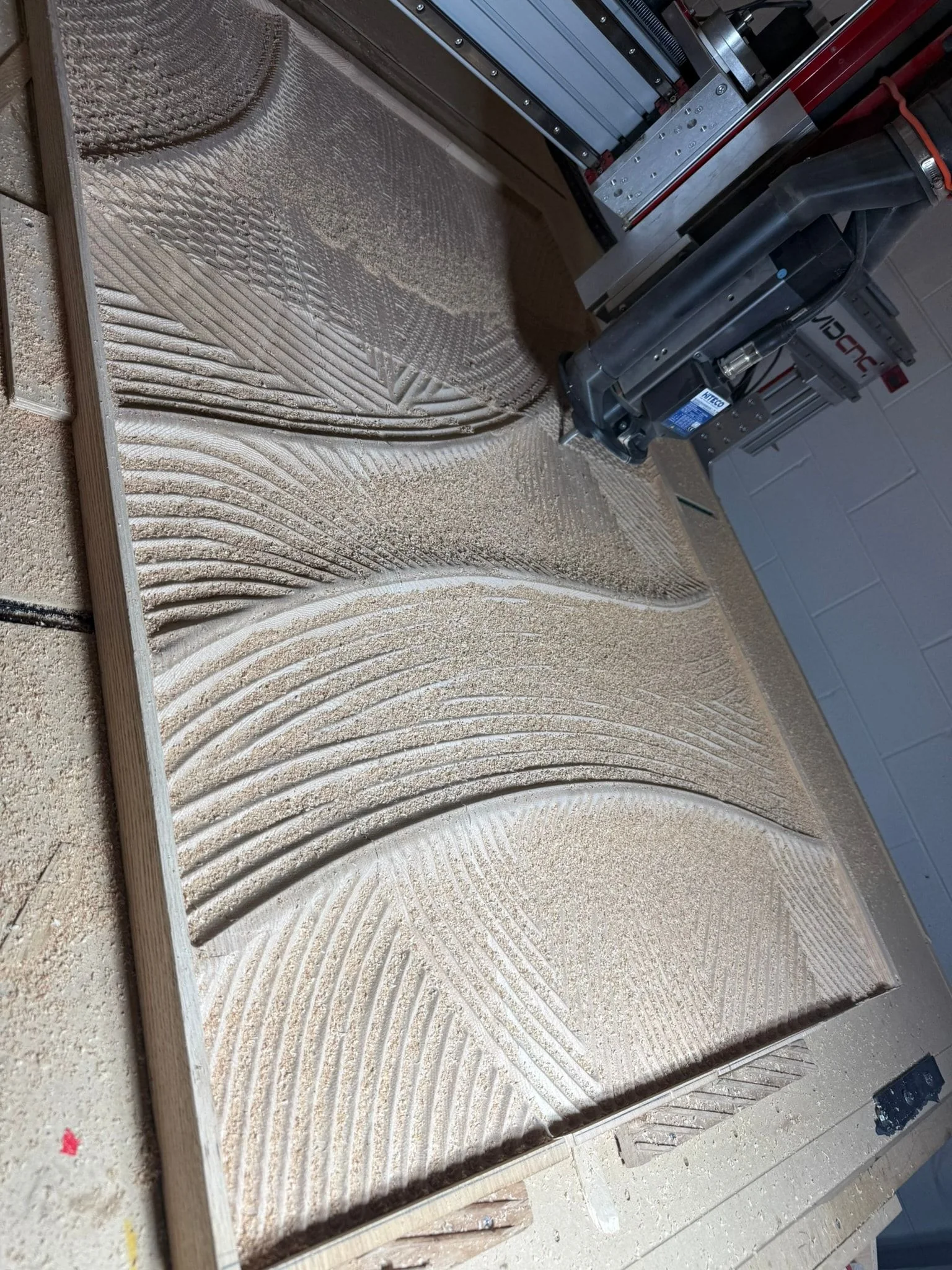 A wave relief half carved into a wood panel on the router bed, covered in fresh dust.