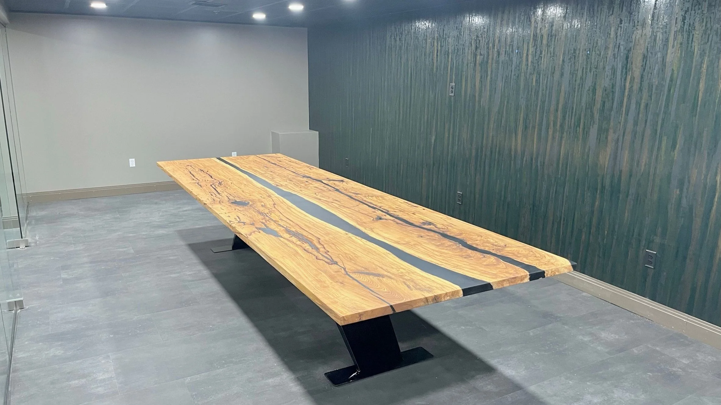 A long conference table with a figured light hardwood top and an angular black steel base, in an office conference room with a dark wall.