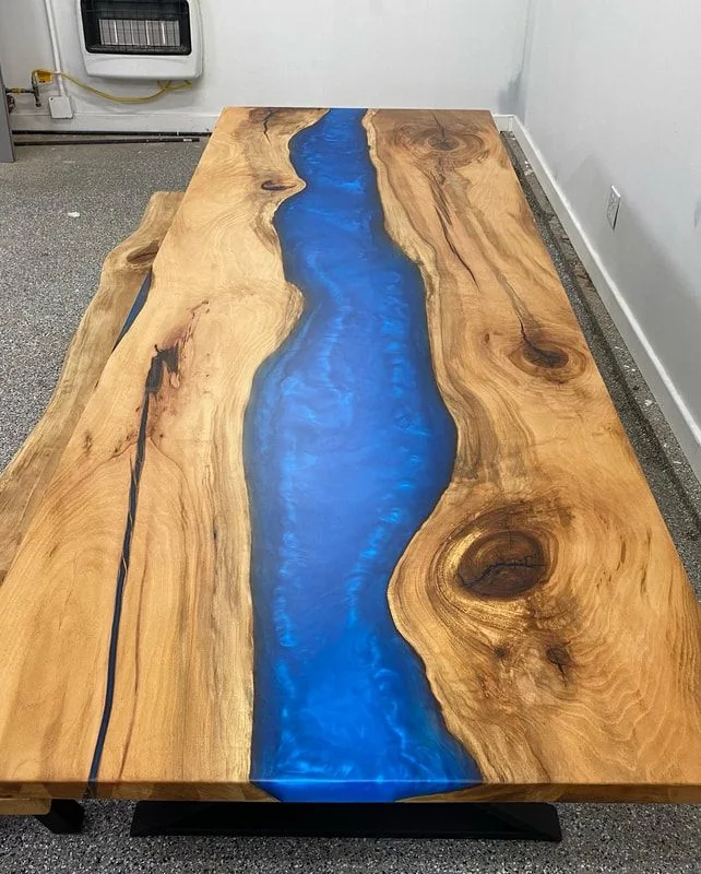 A live edge elm dining table with a broad, deep blue resin river running its full length, on a black base, in the workshop.