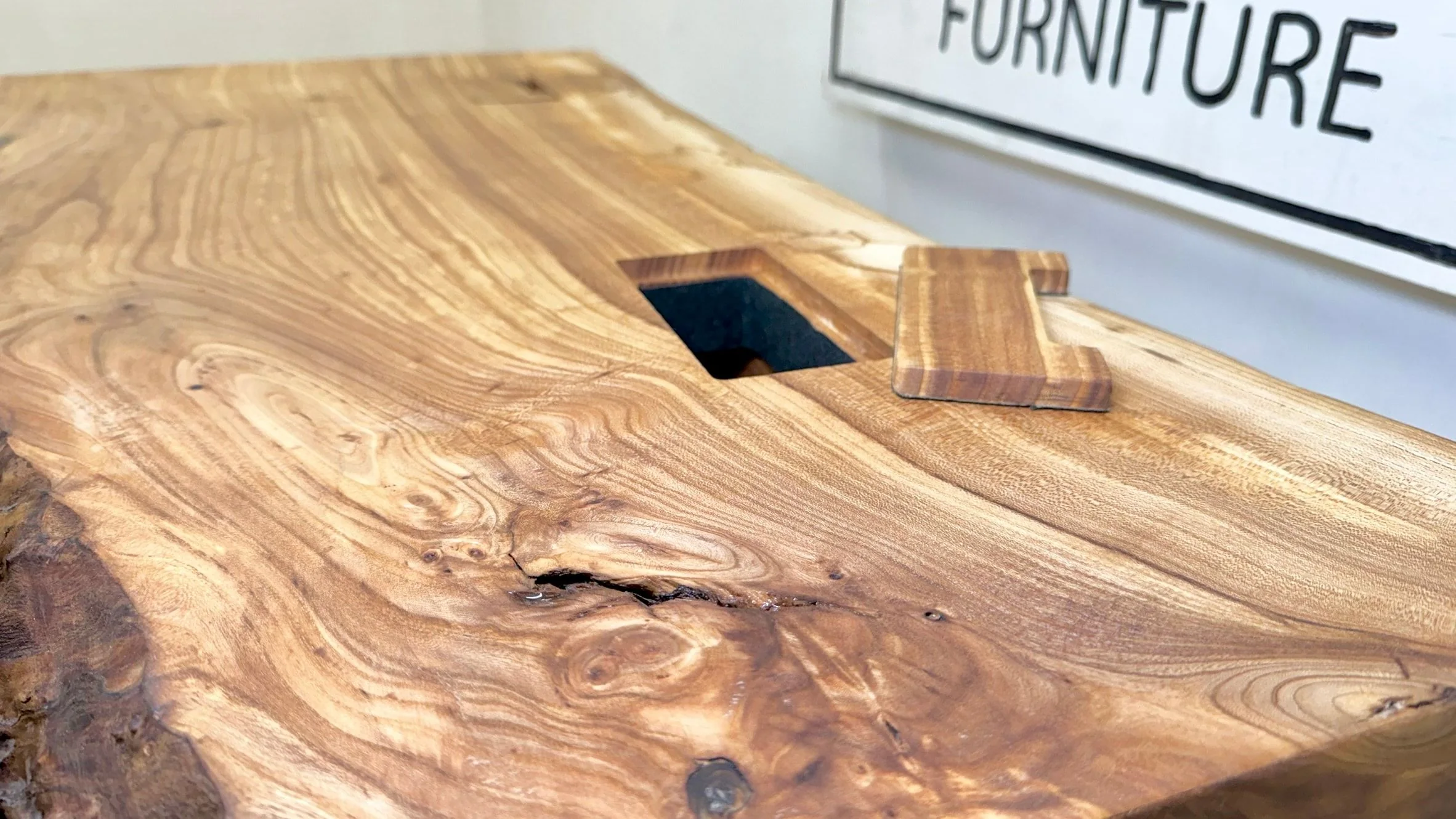 A rectangular port cut through a live-edge desk top, with its fitted wooden cover resting beside it.