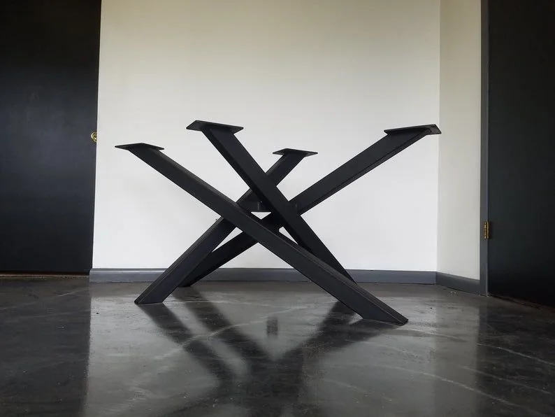 Black metal table base with intersecting crisscross beams forming a geometric support structure, photographed against a dark studio wall.