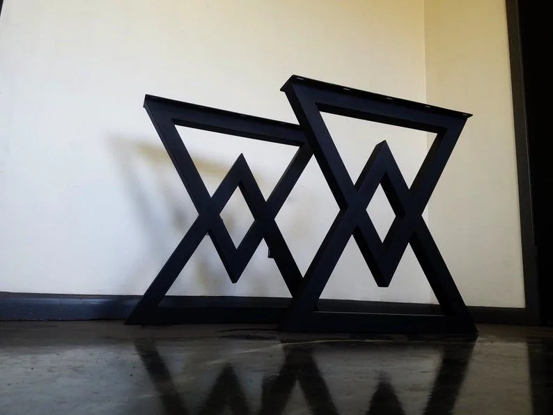 Two black metal table legs with a geometric design featuring a large triangle and an inverted smaller triangle forming a diamond shape, photographed against a dark studio wall.