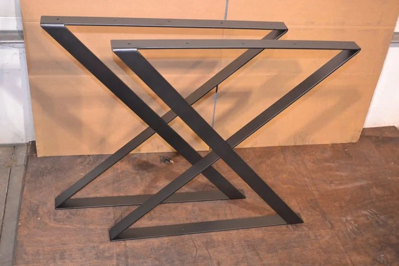 Two black metal table legs in an X-shaped design, placed on a wooden surface with a cardboard background, showcasing a modern and minimalist style.