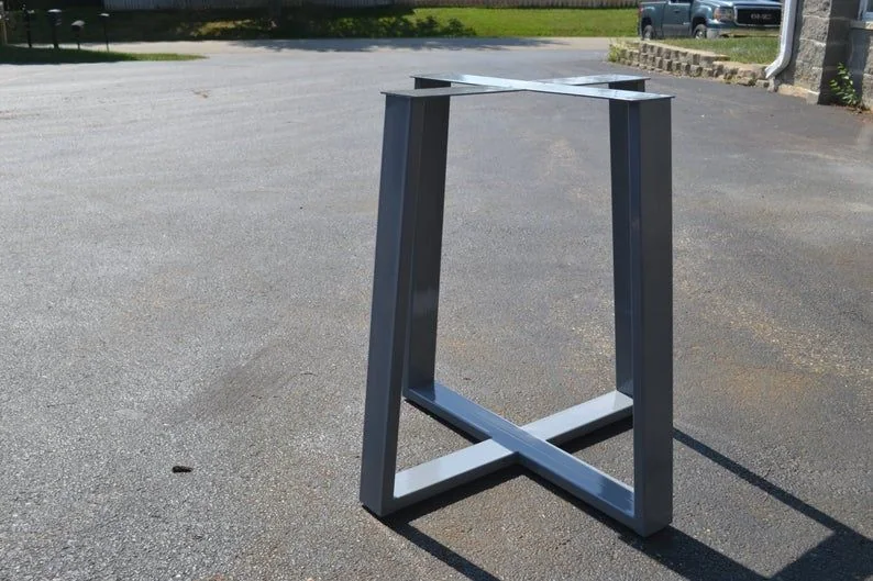 Metal table base with an X-shaped support structure made of rectangular metal beams, placed outdoors on asphalt with greenery and a parked vehicle in the background.
