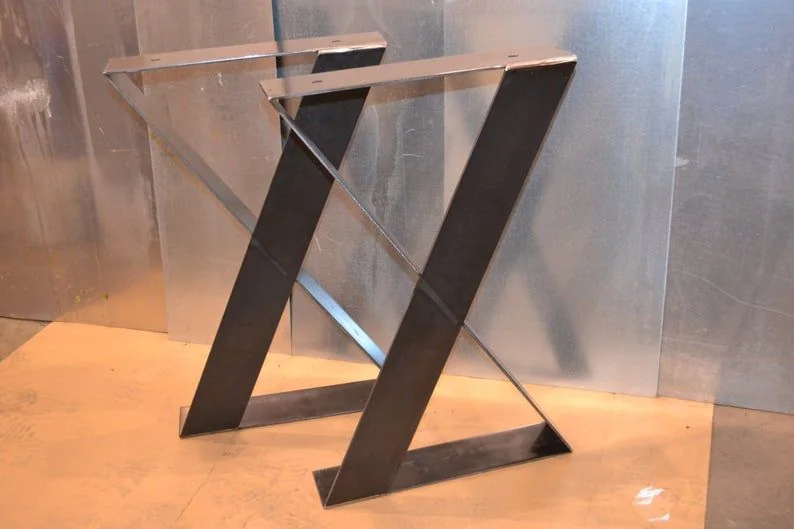 Two steel X-shaped metal table legs with a modern industrial design, placed on a wooden surface against a metallic background.