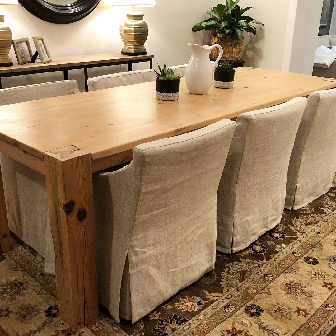Dining room with a large rustic wooden table, six beige slipcovered chairs, decorative plants and pitcher, console table with lamps and framed art, and an oriental rug.