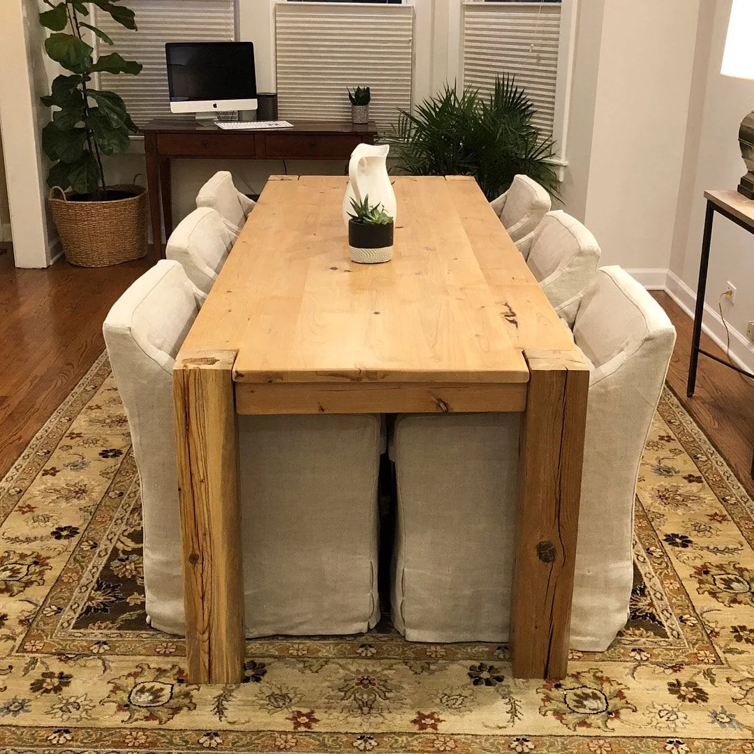 Large rustic wooden dining table with eight cushioned chairs on an ornate rug, decorated with a potted plant and vase, in a bright room with windows and a desk in the background.