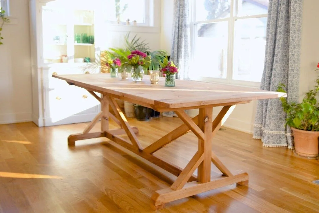 Rustic wooden dining table with X-shaped supports, decorated with vases of flowers, photographed against a dark studio wall.