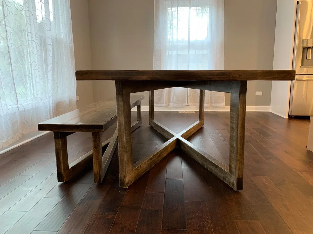 Handcrafted wooden dining table with matching bench featuring X-shaped base, photographed against a dark studio wall.
