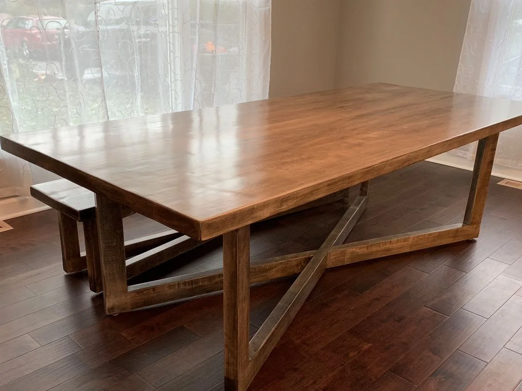 Wooden dining table with a rectangular top and X-shaped support legs, paired with a matching wooden bench, photographed against a dark studio wall.