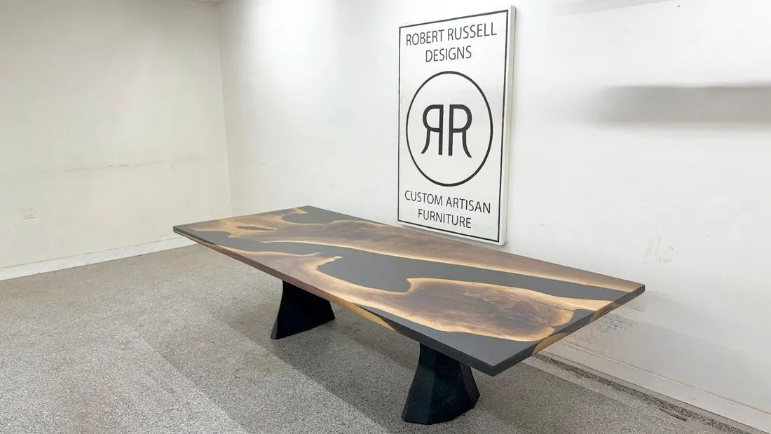 A live edge walnut dining table with a charcoal resin river on two faceted black pedestals.