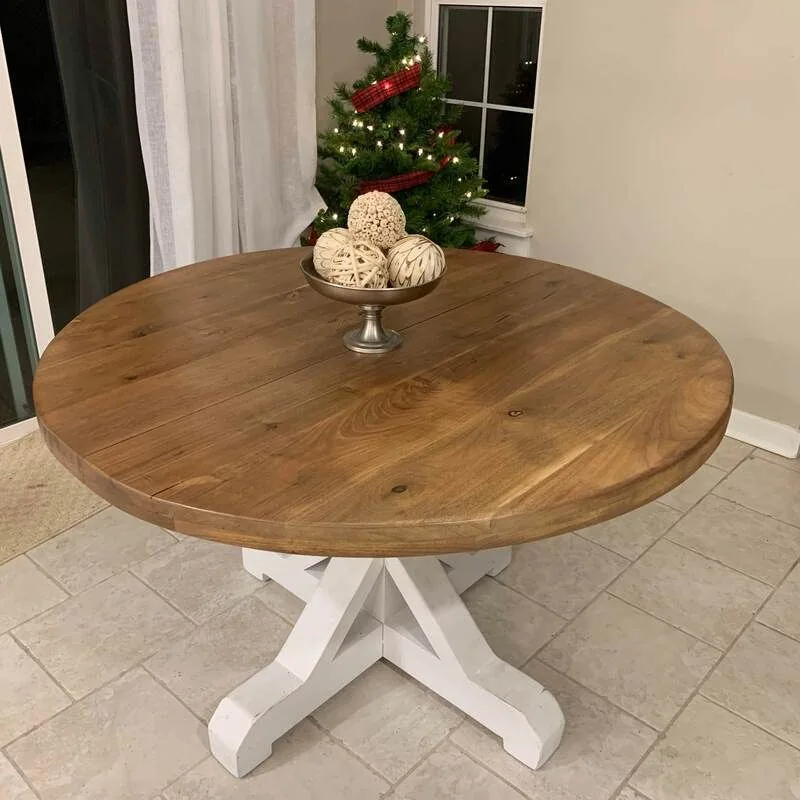 Round wooden table with a white pedestal base, topped with a decorative metal bowl holding assorted spheres, photographed against a dark studio wall.