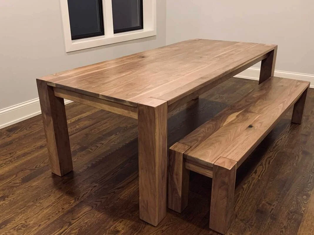 Wooden dining table with matching bench, natural finish, on wooden flooring in a light-colored room.