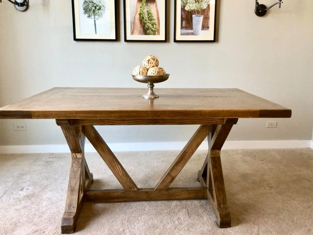 Rustic wooden table with X-shaped supports and a decorative bowl of woven balls, photographed against a dark studio wall.
