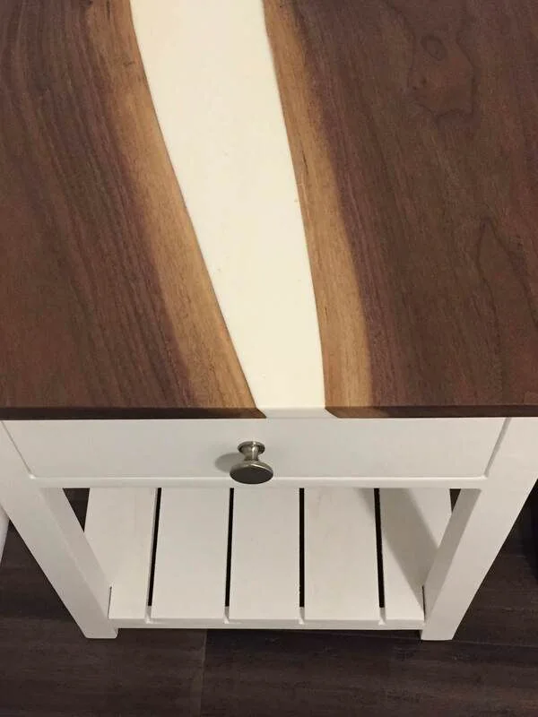 A small wooden nightstand with a white base, a single drawer with a round metallic knob, and a tabletop featuring two dark wood sections flanking a lighter cream-colored center. A slatted shelf is located at the bottom for extra storage.