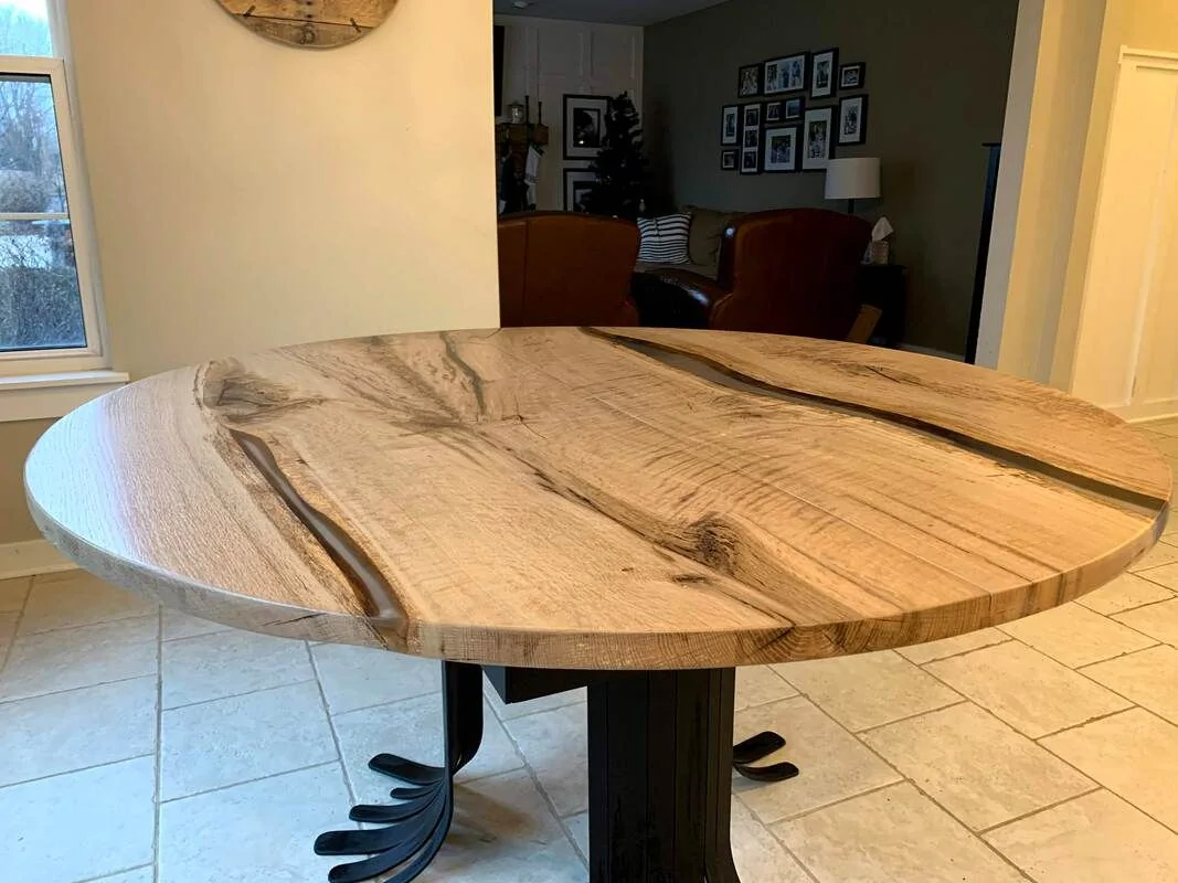 Handcrafted round wooden table with live edge design and black metal base, set in a home interior with tiled flooring and leather chairs.