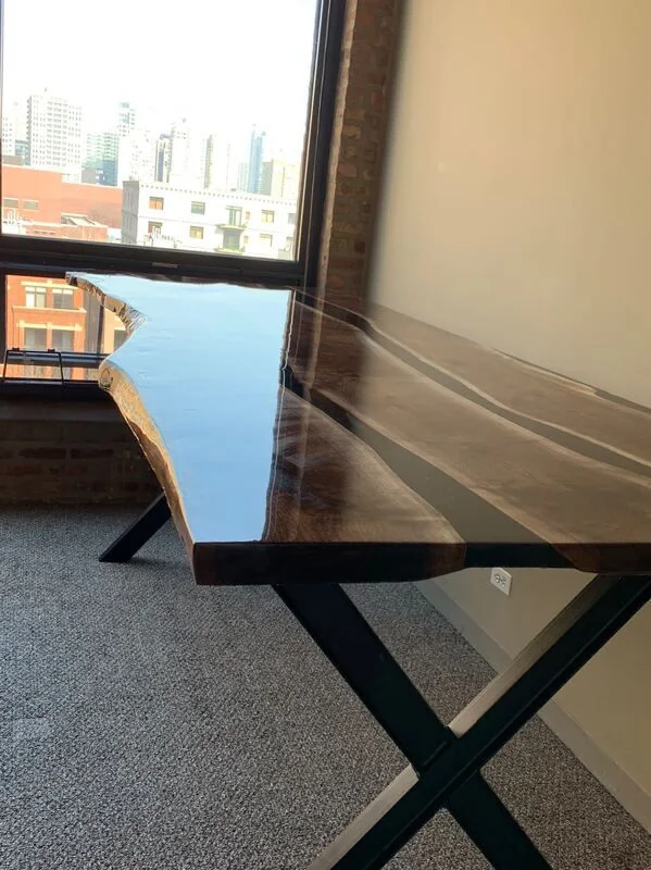 Live edge wood desk with clear resin river and black X-shaped metal legs, photographed against a dark studio wall.
