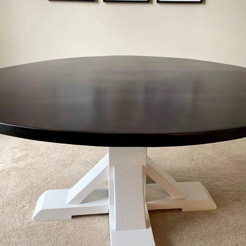 Round table with a dark top and white pedestal base, placed on a carpeted floor beneath three framed pictures on a plain wall