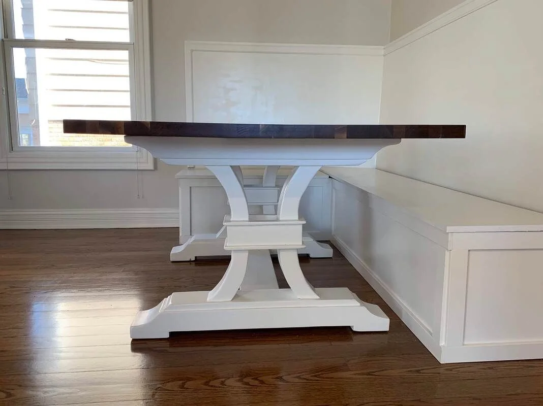 Dark wood dining table with white ornate pedestal legs, paired with built-in white bench seating, photographed against a dark studio wall.