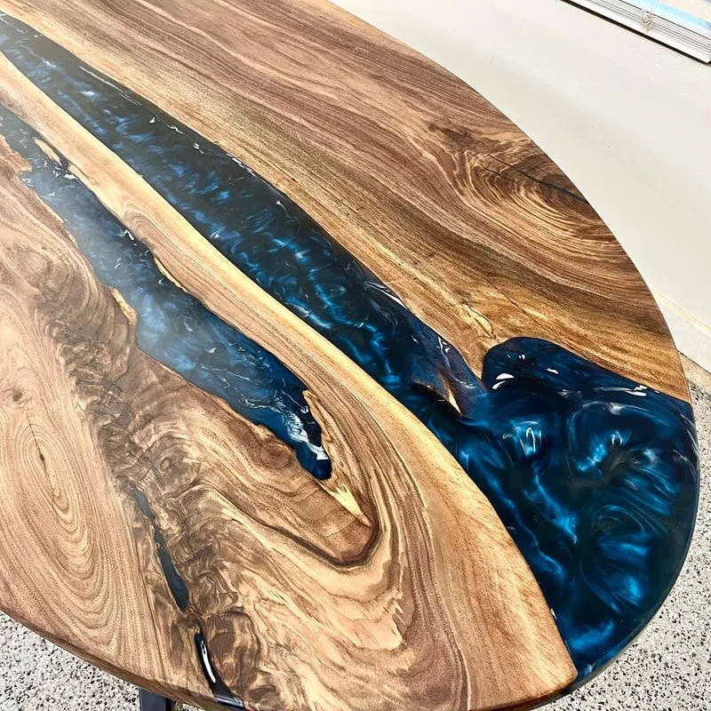 Close view of an oval walnut table top, a deep blue resin river widening into a pool between heavily figured live edges.
