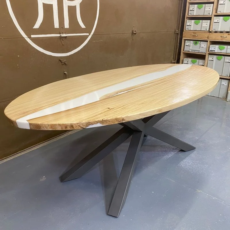 An oval table in pale ash with a white resin river, on a grey steel spider base, photographed against a dark studio wall.