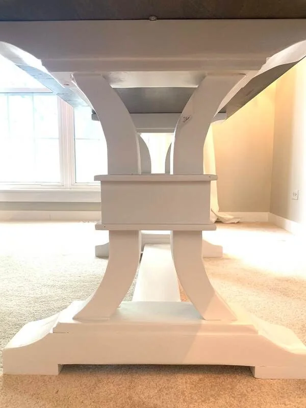 Underside of a table with white curved supports and a horizontal connector, photographed against a dark studio wall.