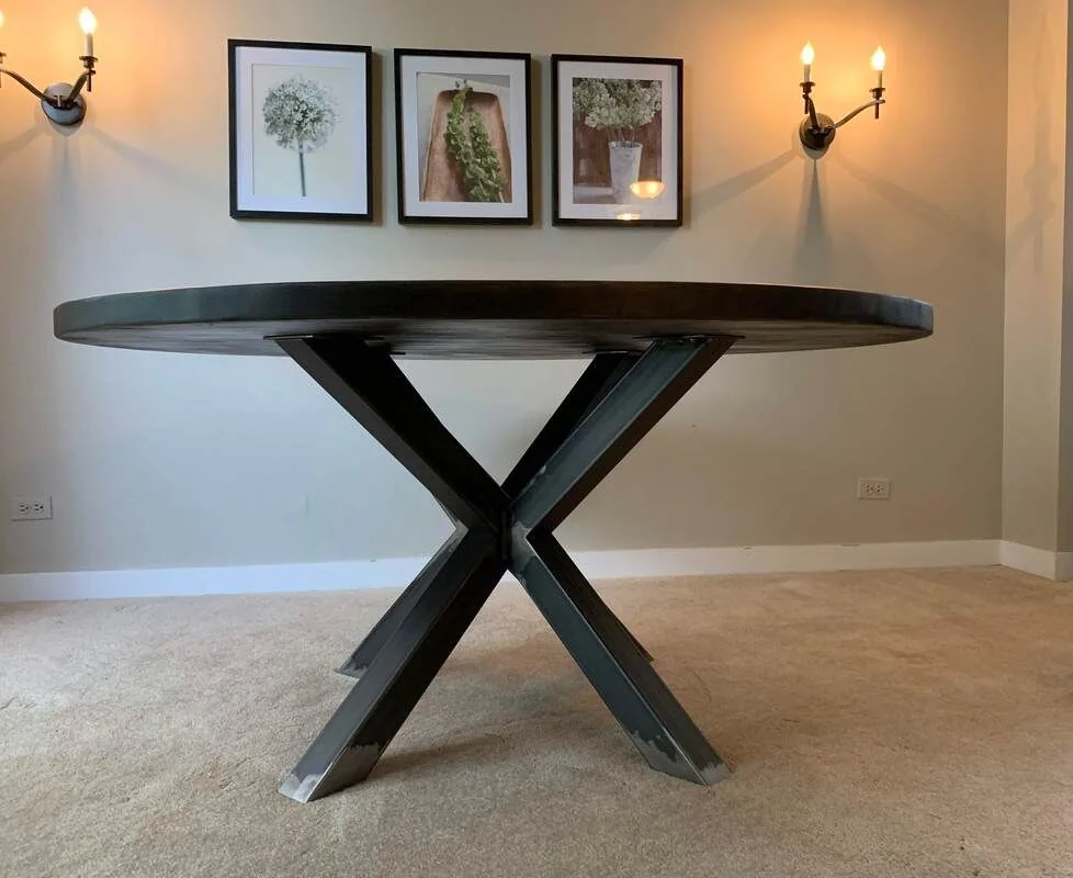 A round table with a thick, dark wood top and a black X-shaped metal base, set on a carpeted floor in a room with light-colored walls. Behind the table are three framed floral prints and two wall-mounted candle sconces with lit candles, creating a cozy and decorative interior setting.