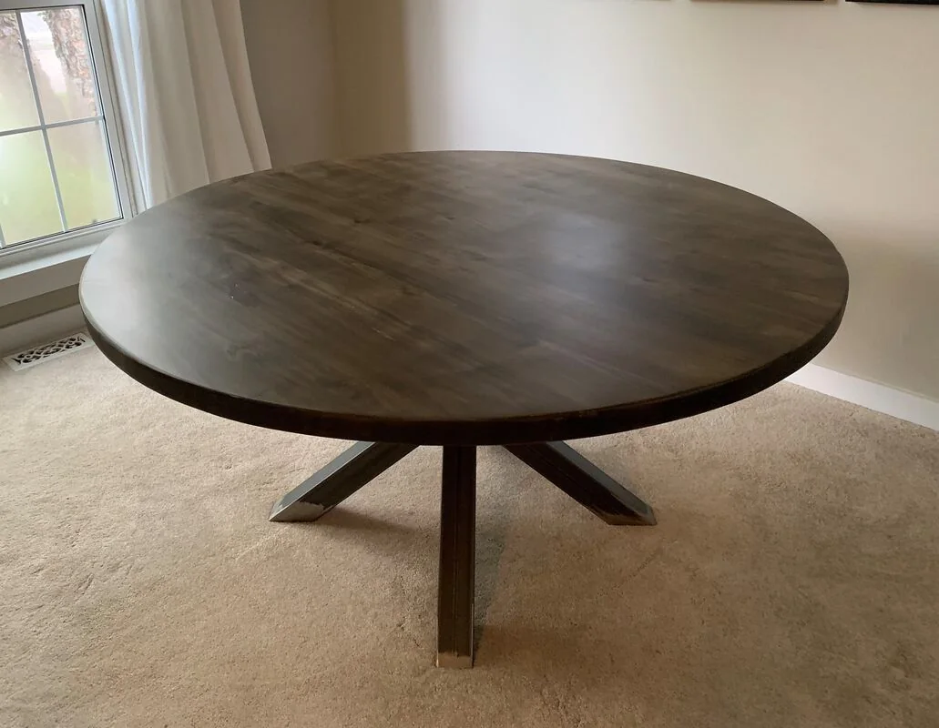 Round wooden table with a dark finish and pedestal base on a carpeted floor near a window with white curtains.