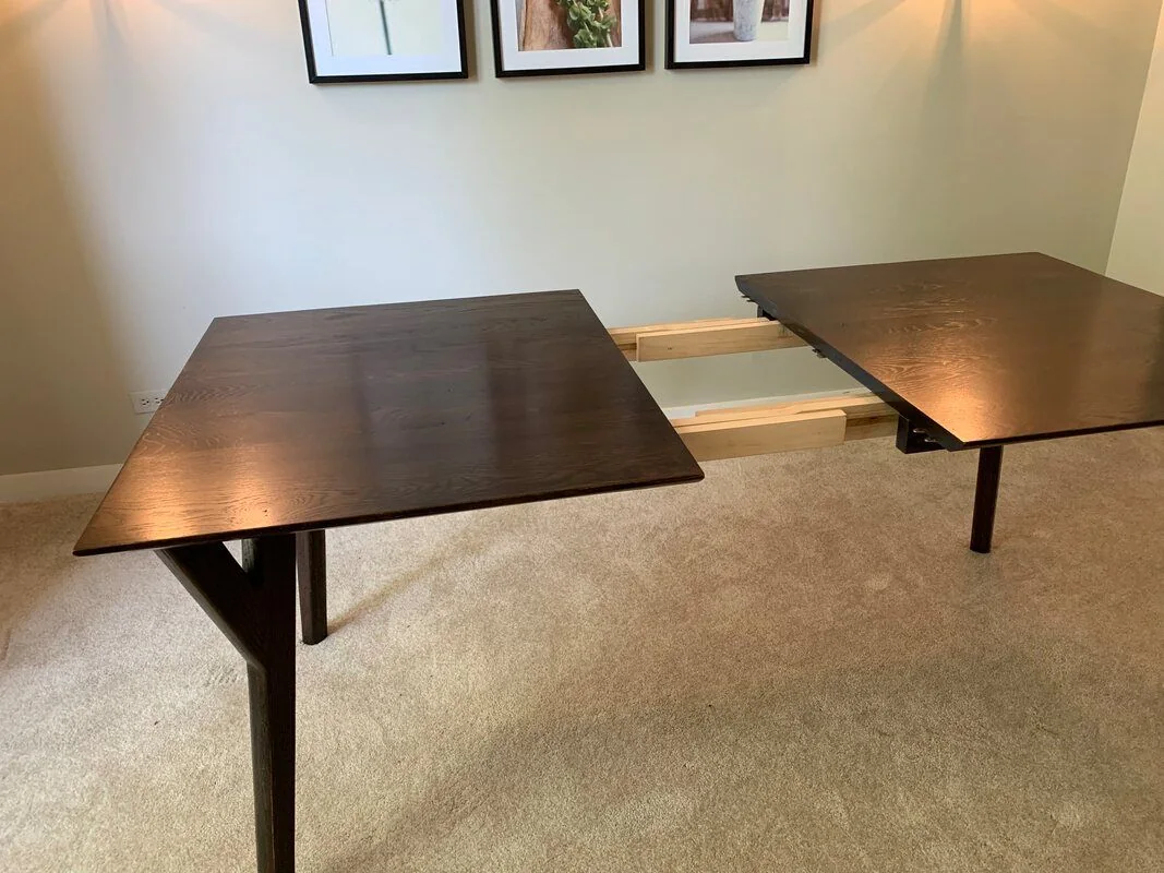 Dark wood extendable dining table split in the middle for leaf insertion, placed on carpeted flooring with framed wall art and wall-mounted lights in the background.