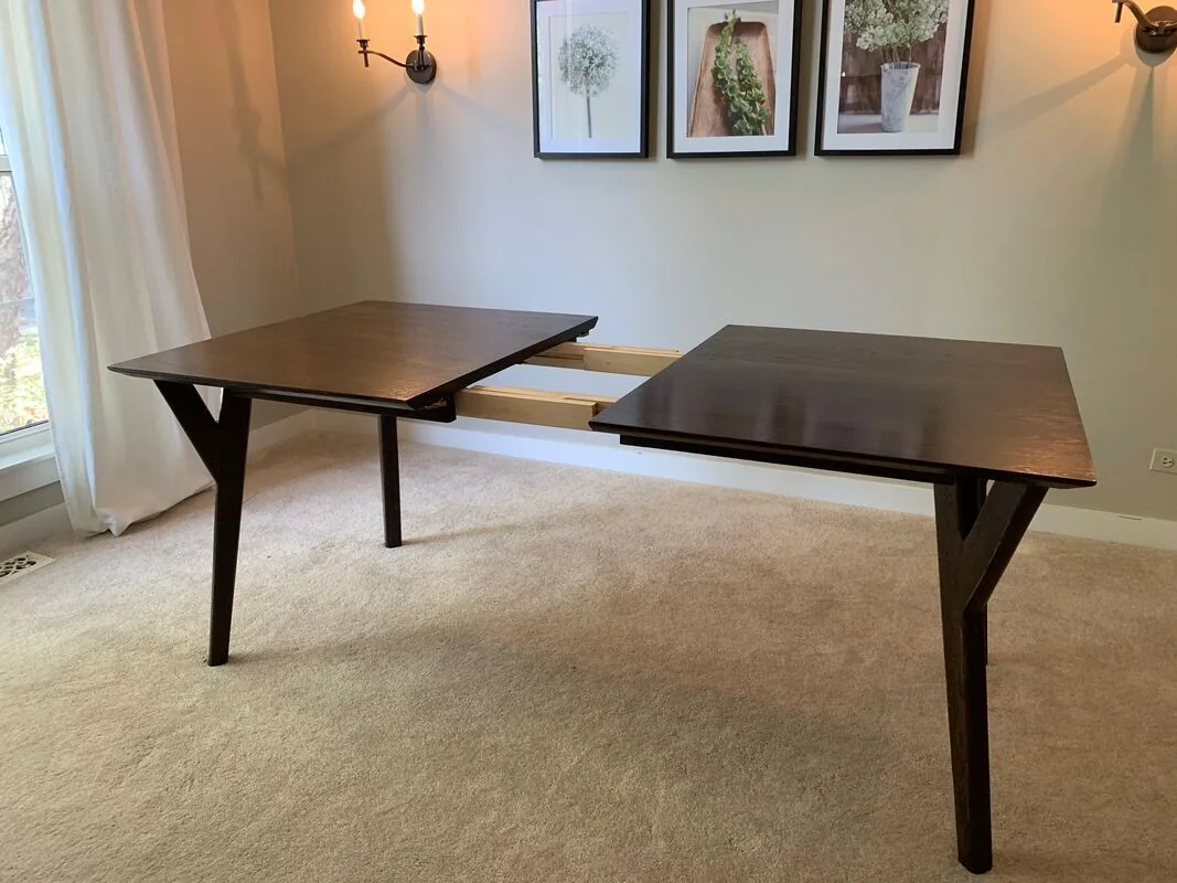Dark wood dining table shown partially extended with a center gap for leaf insertion, placed on carpeted flooring with framed artwork and wall-mounted lights on the wall behind.