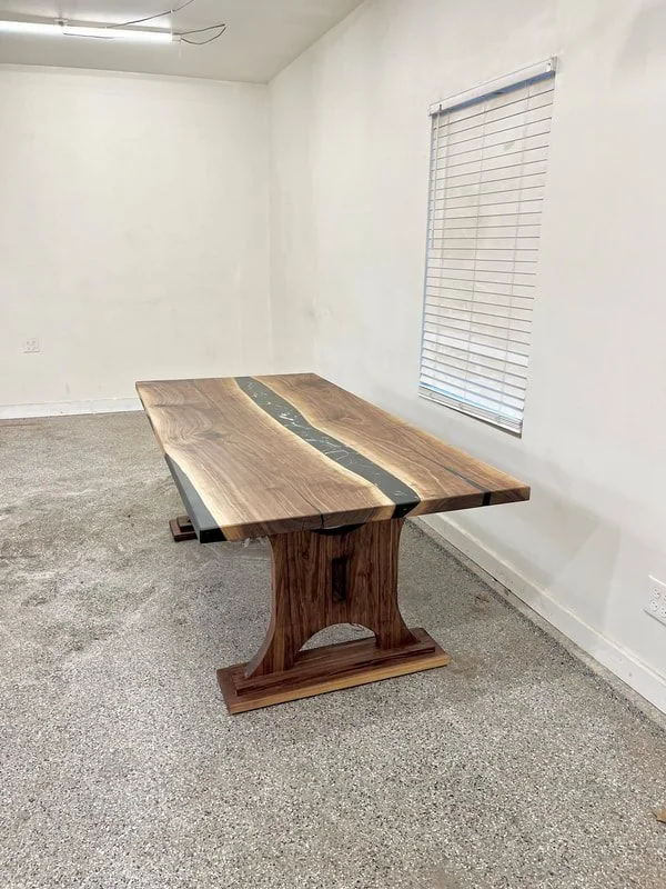 Wooden table with two slabs joined by a dark resin river, featuring sturdy wooden legs, photographed against a dark studio wall.