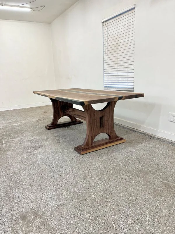 Solid wood table with natural grain, thick curved legs, and a horizontal support beam, placed in a room with white walls and gray speckled flooring.