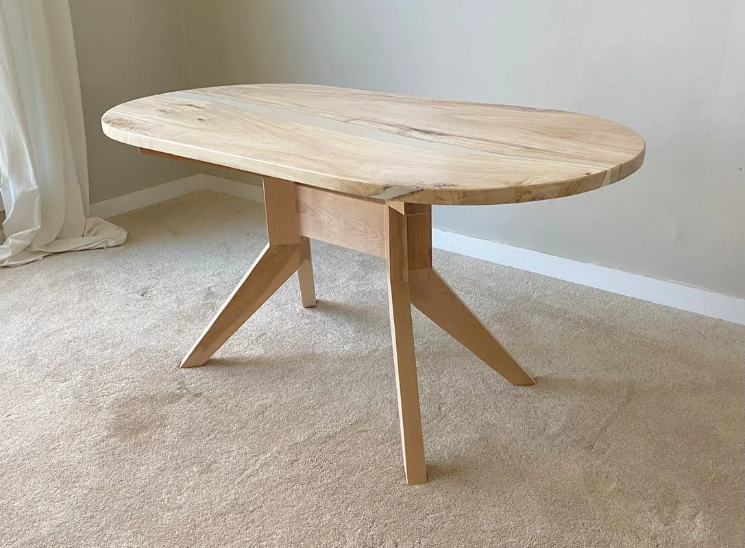 An oval maple table with a thin pale resin line across the top, on a splayed four-leg maple base, unfinished, photographed against a dark studio wall.