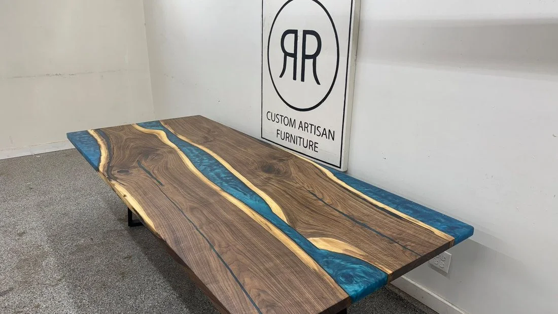 A live edge walnut dining table with two bright blue resin rivers on black steel legs, photographed against a dark studio wall.