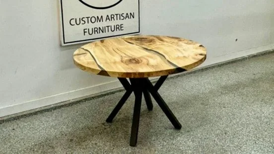A small round table in pale figured wood with a clear resin river, on a black spider base, photographed against a dark studio wall.