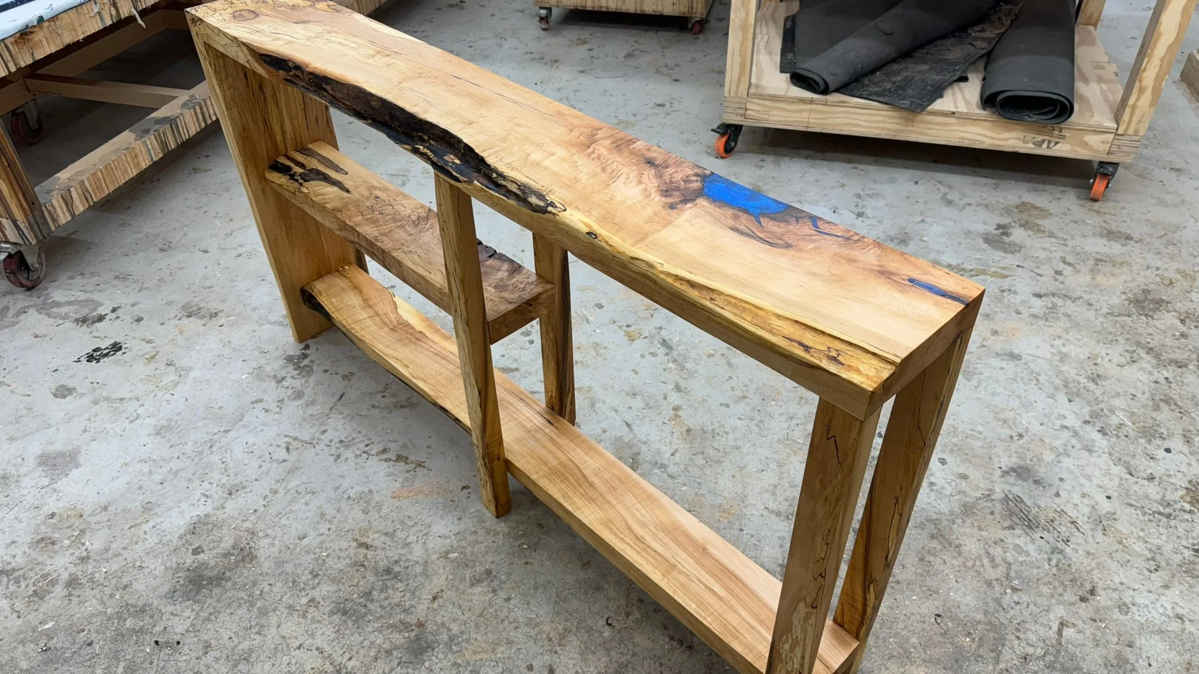 A live-edge console table with a blue resin fill, standing on the concrete floor of the studio before its finish.