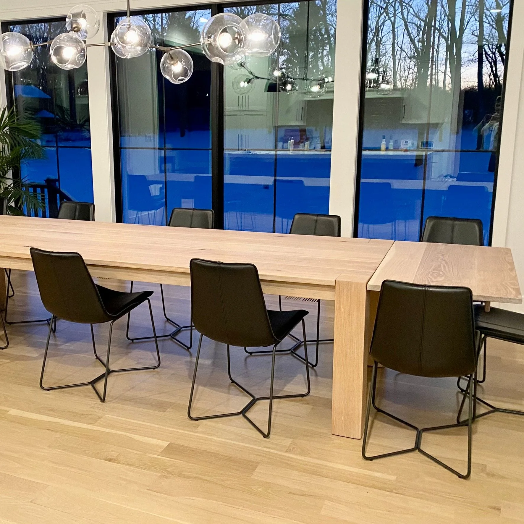 A long pale hardwood dining table on thick square legs, black chairs along it and floor-to-ceiling windows behind, installed in a client’s home at dusk.