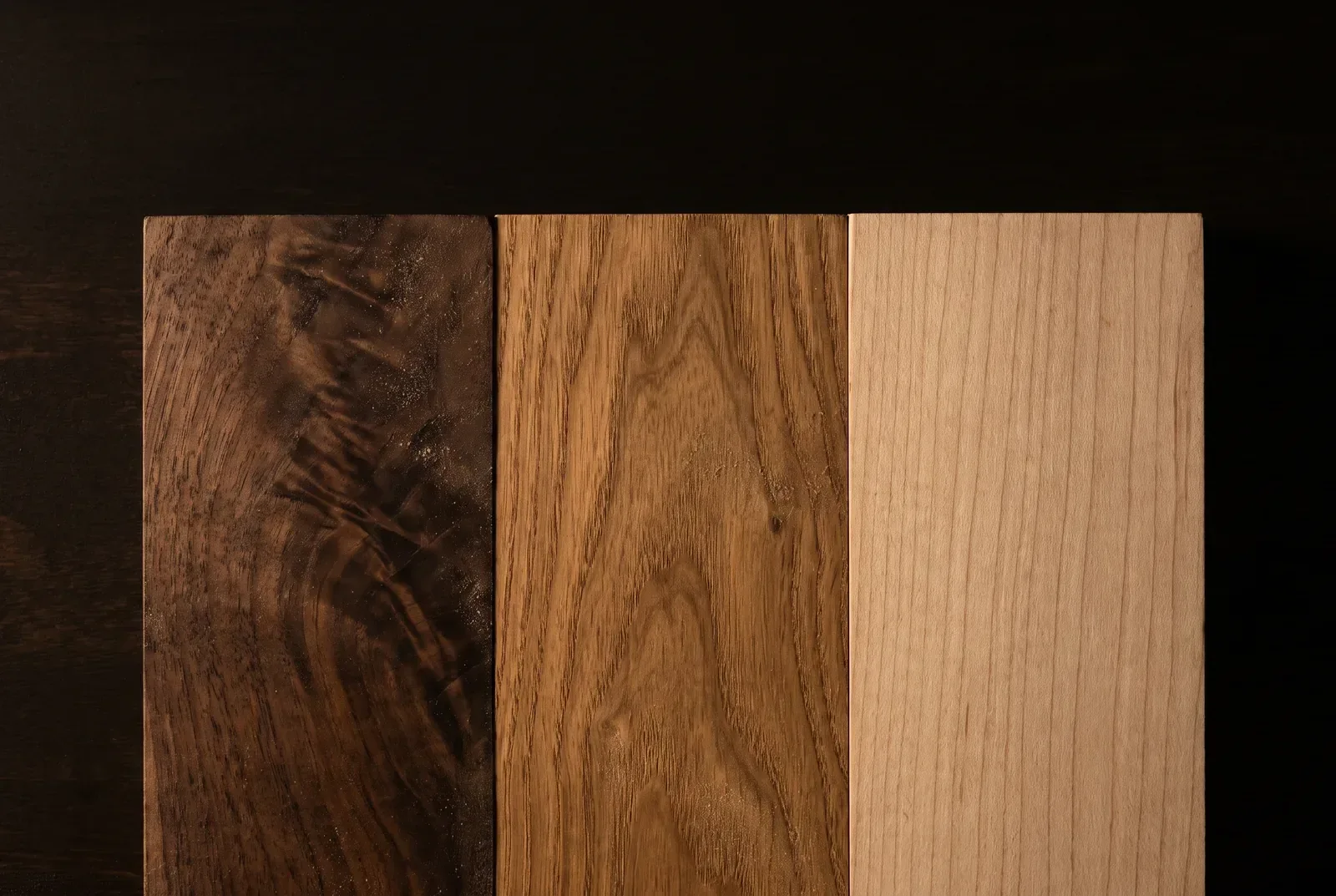 Three hardwood samples laid side by side on a dark bench: dark figured walnut, warm mid-brown elm and pale straight-grained maple.