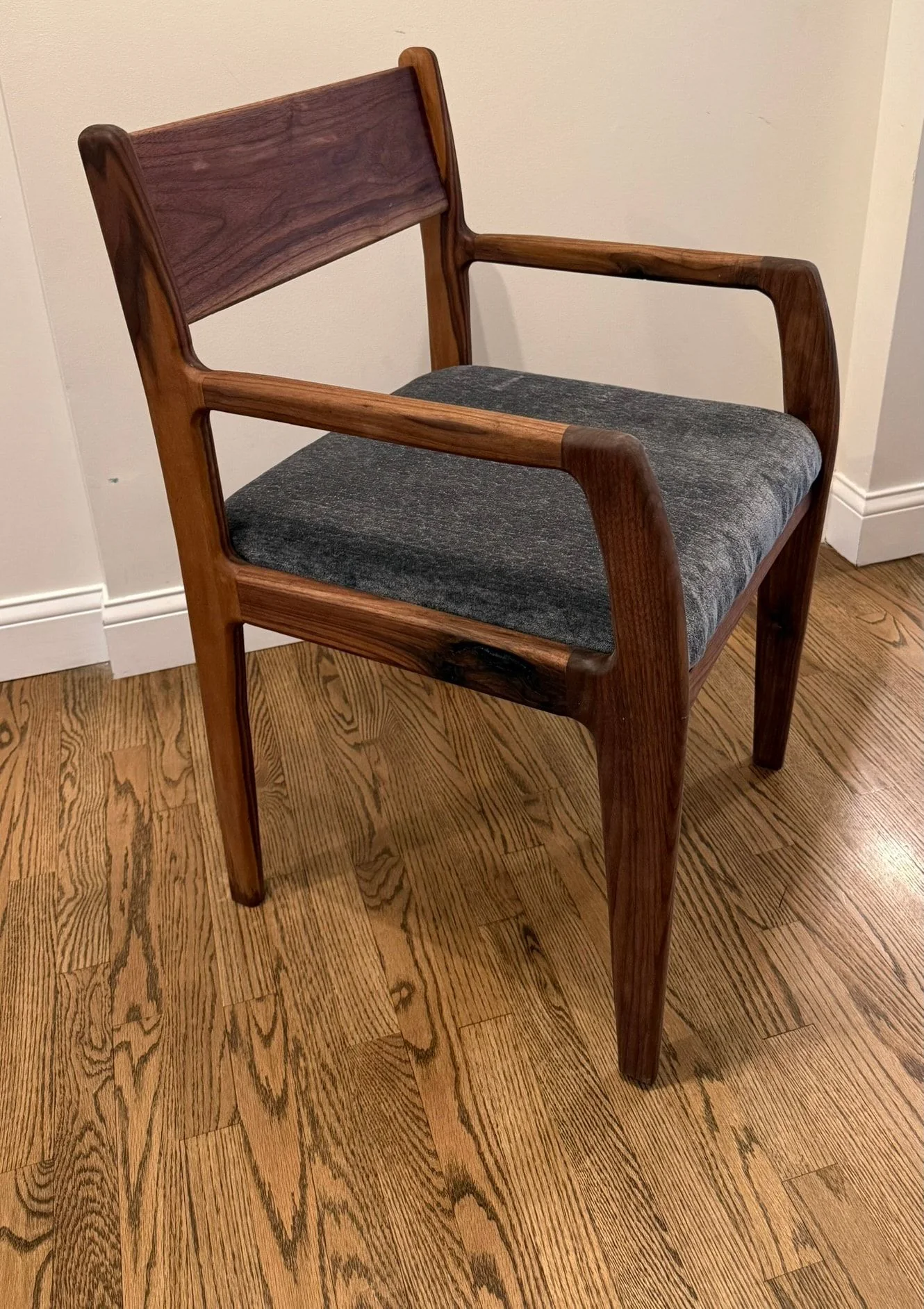 A walnut armchair with shaped arms, a solid back rail and a grey upholstered seat, on a hardwood floor.
