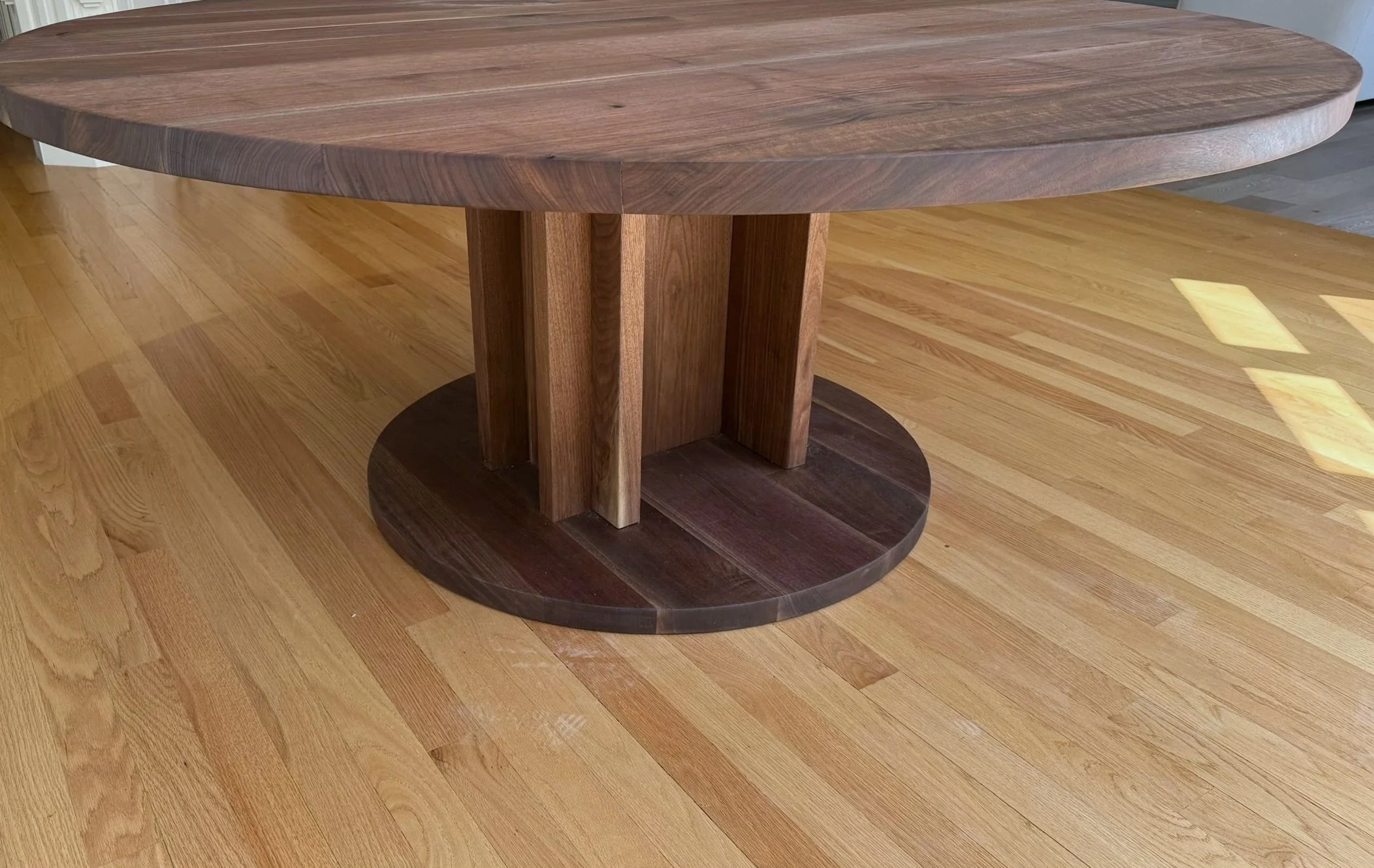 The same table built in walnut: a round top on a pedestal of vertical boards set in a cross, on a round walnut plinth, on a hardwood floor.