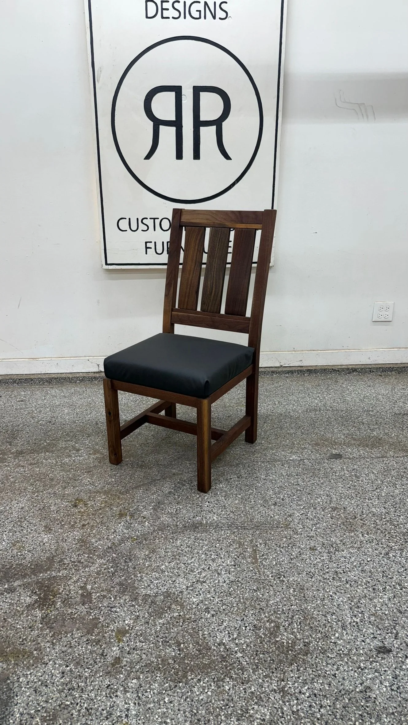 A walnut dining chair with a tall slatted back and a black upholstered seat, in the workshop.