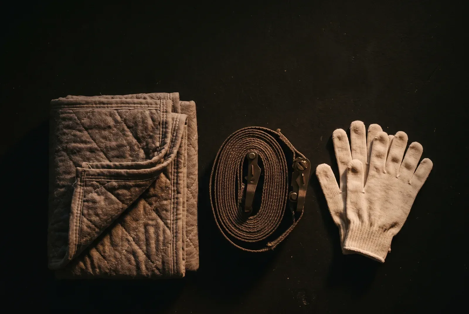 A folded quilted moving blanket, a coiled ratchet strap and a pair of white cotton gloves laid on a dark floor.