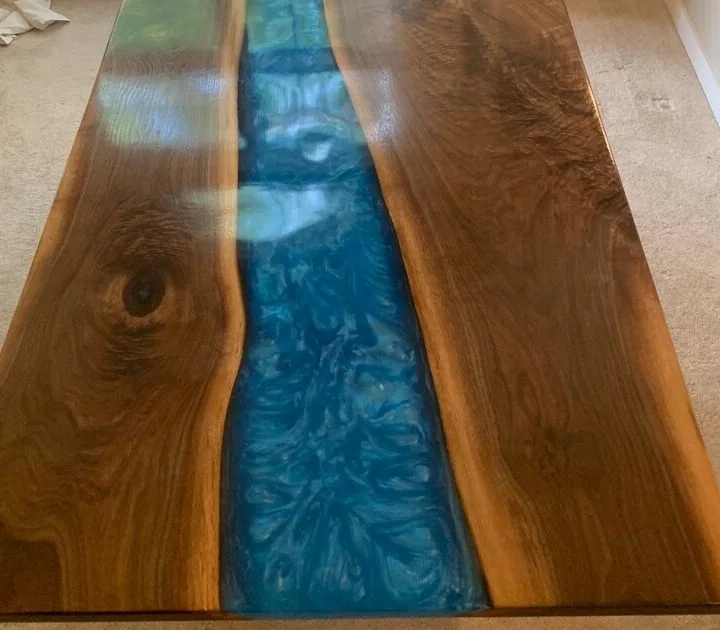 A wooden table with two natural-edge slabs joined by a marbled blue resin river, placed on beige carpet near a window overlooking a green lawn and outdoor seating area.