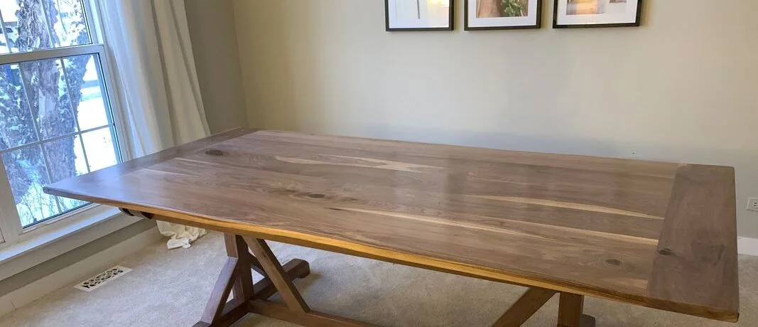 Large wooden dining table with X-shaped supports on beige carpet, near a wall with botanical prints and light fixtures.