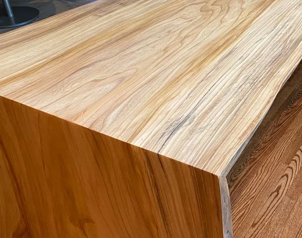 Large live edge wood slab table with polished surface and natural grain, set indoors with wood flooring and modern bar stools.